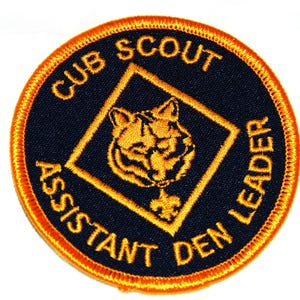 May include: A circular patch with a navy blue background and orange embroidered border. The patch features the text "CUB SCOUT ASSISTANT DEN LEADER" in orange. A gold-colored wolf head is inside a gold diamond.