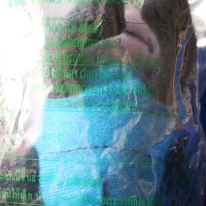 May include: A blue and green plush toy in a clear plastic bag. The toy is wrapped in a blue and green fabric. The bag has text in multiple languages, including English, Japanese, and Arabic. The text includes the words "Supplier: NA PROMOTION (ASIA)" and "Admiralty, Hong Kong".