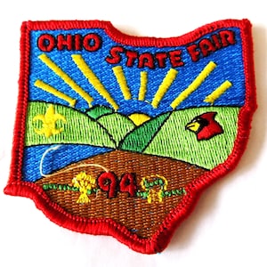 May include: Embroidered patch in the shape of Ohio with the words "Ohio State Fair" in red. The patch features a blue sky with yellow sun rays, green hills, and a red cardinal. The year "94" is also displayed.