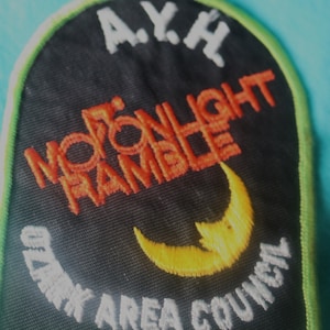 May include: Black and white embroidered patch with a crescent moon and the text "Moonlight Ramble" in orange. The text "A.Y.H. Ozark Area Council" is also embroidered in white.