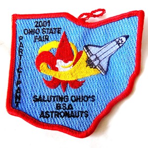 May include: Embroidered patch in the shape of Ohio with a red border. The patch features a blue background with text and images including a space shuttle, a fleur-de-lis, and the words "2001 Ohio State Fair".