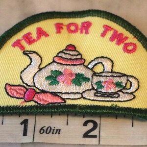 May include: Embroidered patch with a yellow background and green border. It features a teapot and teacup with pink flowers and the text "Tea for Two".