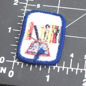 May include: A blue and white embroidered patch with a red, yellow, and black design. The design features a group of people standing on a platform with a blue ribbon.
