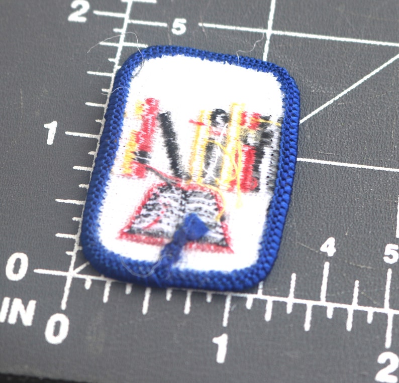 May include: A blue and white embroidered patch with a red, yellow, and black design. The design features a group of people standing on a platform with a blue ribbon.