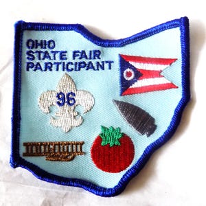 May include: Embroidered patch in the shape of Ohio, light blue background with a dark blue border. Features text "OHIO STATE FAIR PARTICIPANT", a fleur-de-lis with "96", Ohio flag, tomato, and arrowhead.