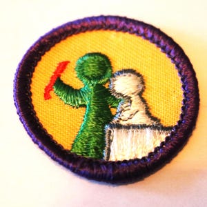 May include: A purple and yellow embroidered Girl Scout badge featuring a green stick figure holding a red stick and a white stick figure standing on a white box.