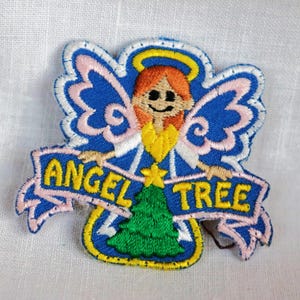 May include: An embroidered patch featuring an angel with a halo, pink wings, and a yellow dress. The angel is holding a green Christmas tree with a yellow star on top. The text "ANGEL TREE" is written on a blue banner.
