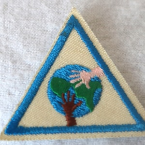 May include: A triangular Girl Scout patch with a blue border. The patch features a white background with a brown tree and two hands reaching towards a blue globe with green continents.