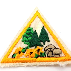 May include: Triangular patch with an embroidered design featuring green trees, yellow flowers, and brown mushrooms. The patch has a yellow border and is set against a cream-colored background. The patch is likely for a scout or similar organization.