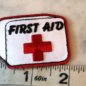 May include: White fabric patch with red embroidered cross and black embroidered text "FIRST AID". The patch is approximately 1 1/2 inches wide.