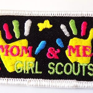 K&ouml;nnte beinhalten: Gestickter Aufn&auml;her mit den Worten "MOM & ME GIRL SCOUTS" in Gelb und Gr&uuml;n. Der Aufn&auml;her zeigt zwei gelbe Handabdr&uuml;cke mit bunten Fingerspitzen, einen rosa Schmetterling und einen gelben Stern auf schwarzem Hintergrund mit wei&szlig;em Rand.