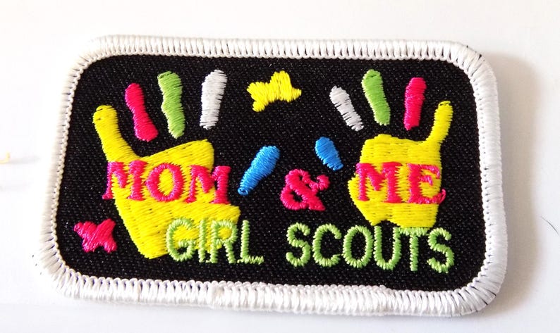 K&ouml;nnte beinhalten: Gestickter Aufn&auml;her mit den Worten "MOM & ME GIRL SCOUTS" in Gelb und Gr&uuml;n. Der Aufn&auml;her zeigt zwei gelbe Handabdr&uuml;cke mit bunten Fingerspitzen, einen rosa Schmetterling und einen gelben Stern auf schwarzem Hintergrund mit wei&szlig;em Rand.