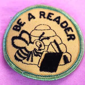 May include: A yellow and green embroidered patch with a bee reading a book. The text on the patch reads "Be a Reader".