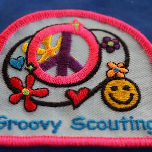 May include: A blue and pink embroidered patch with a peace sign, flowers, a smiley face, and the text "Groovy Scouting".