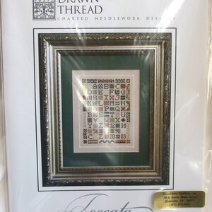 May include: A white and brown cross stitch pattern with the alphabet and numbers in a frame. The pattern is titled "Loccata Number One" and is from The Drawn Thread Charted Needlework Designs.
