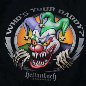 May include: A black t-shirt with a graphic of a scary clown with a green and purple hat. The clown is holding a bone in each hand and has a large, toothy grin. The text "Who's Your Daddy?" is written in a circle around the clown. The text "Hellaback Incorporated" is written below the clown.