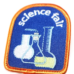 May include: A blue patch with the words "science fair" in white, arched across the top. Two lab flasks are depicted in white and light blue, with yellow liquid inside. The patch has an orange border.