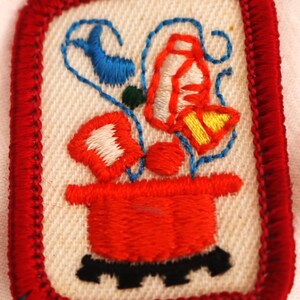 May include: Embroidered patch featuring a red pot with flowers in red, white, and yellow. The patch has a red border and a cream-colored background. Blue and green accents add detail to the floral design.