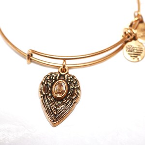 May include: Gold charm bracelet with a heart-shaped charm featuring angel wings and a clear crystal. The bracelet has two additional charms, one with a star design and the other with an American flag design.