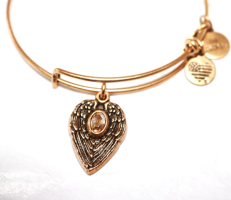 May include: Gold charm bracelet with a heart-shaped charm featuring angel wings and a clear crystal. The bracelet has two additional charms, one with a star design and the other with an American flag design.