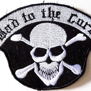 May include: Embroidered patch featuring a skull with crossed bones, set against a black background. The patch has a white border with the words "Bad to the Core" in a gothic font. The skull and bones are white.