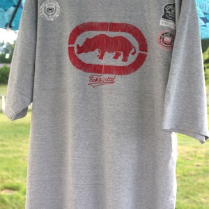 May include: Gray t-shirt with a red rhinoceros logo and the text "Ecko Unltd." The shirt has several small patches with logos.