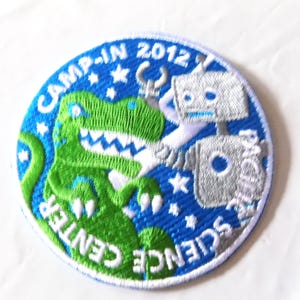 May include: Embroidered patch with a blue background featuring a green dinosaur and a silver robot. The text "CAMP-IN 2012" and "PACIFIC SCIENCE CENTER" are in white. The patch is round.