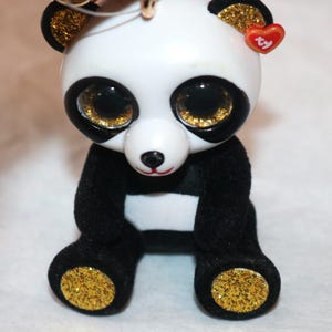 May include: A black and white panda bear plush keychain with gold glitter accents. The panda has large, black eyes with gold glitter pupils and a red Ty heart tag.