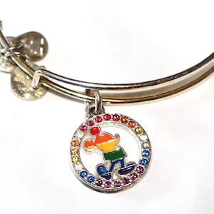 May include: Silver-toned bangle bracelet with a round charm featuring a colorful cartoon character. The charm is encircled by a ring of multi-colored gemstones. The bracelet has a simple, elegant design.