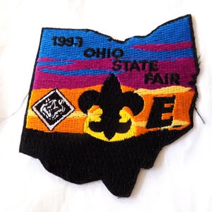 May include: Embroidered patch in the shape of Ohio, featuring text "1993 OHIO STATE FAIR". The patch has a black fleur-de-lis, a diamond emblem, and a colorful sunset design in blue, purple, orange, and yellow.