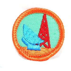 May include: A round, orange-bordered patch with a green background. The patch features a blue and red embroidered image of a radio tower and a satellite dish.