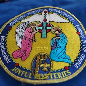 May include: A round, yellow embroidered patch with a gold border. The patch features a white dove with a red cross and a rosary. Two angels are on either side of the cross, one in pink and one in blue. The text around the border reads "Visitation Nativity Presentation Finding in the Temple Annunciation Joyful Mysteries".
