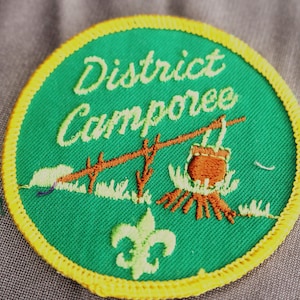 May include: A round, embroidered patch with a green background and yellow border. The text "District Camporee" is in white script. A campfire scene with a pot hanging over it is in the center. A fleur-de-lis is at the bottom.