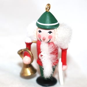 May include: A wooden nutcracker ornament with a red and white coat, a green hat, and a white fur trim. The nutcracker is holding a gold trumpet.