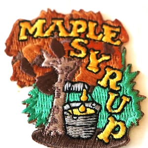 May include: Embroidered patch featuring the words "MAPLE SYRUP" in yellow, with a tree and a bucket design. The patch has brown, orange, green, and yellow colors. The patch is likely for clothing or accessories.