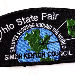 May include: Black embroidered patch with the words "Ohio State Fair" and "Salutes Scouting Around the World." Features a green outline of Ohio and a blue globe. Includes the text "Simon Kenton Council" and the year "1999."