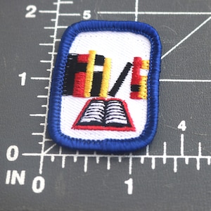 May include: Blue and white embroidered patch with a red, yellow, and black book stack and an open book with white pages.