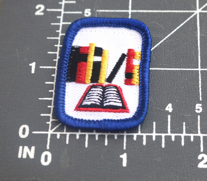 May include: Blue and white embroidered patch with a red, yellow, and black book stack and an open book with white pages.
