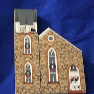 May include: A wooden church building with a gray roof and stone walls. The church has three windows with wreaths and a double door with a wreath. The church is decorated for Christmas.