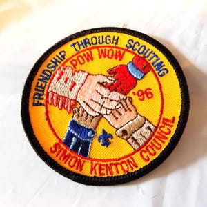 May include: Embroidered patch with a yellow background and black border. The patch features the text "Friendship Through Scouting Pow Wow '96 Simon Kenton Council" around the edge. Two hands are clasped in the center, with red, blue, and brown accents.