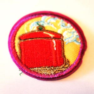 Puede incluir: Un parche redondo bordado de color rosa y amarillo con una olla roja con vapor saliendo de ella.