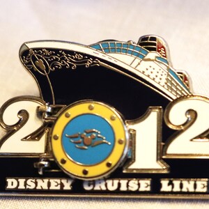May include: A Disney Cruise Line pin from 2012, featuring a cruise ship design in navy blue, white, and blue. The pin includes the text "2012" and "Disney Cruise Line" in white lettering on a black background. A Mickey Mouse icon is in the center.