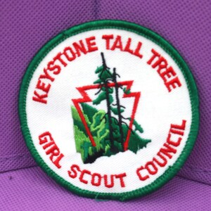 May include: A white, green, and red embroidered patch with the text "Keystone Tall Tree Girl Scout Council". The patch features a stylized image of a tree and a mountain range.