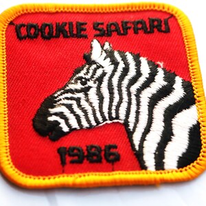 May include: Embroidered patch with a red background, featuring a zebra head in black and white. The text "COOKIE SAFARI" is above the zebra, and "1986" is below. The patch has a yellow border.