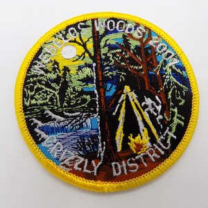 May include: Embroidered patch with a yellow border featuring a forest scene with a river, trees, and a campfire. Text includes "WEBELOS WOODS 2007" and "GRIZZLY DISTRICT". The patch is circular.