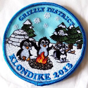 May include: Embroidered patch with a light blue background and a blue stitched border. The patch features three penguins around a campfire, with the text "Grizzly District" and "Klondike 2013". Snow and trees are also depicted.