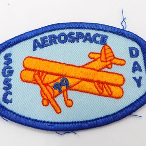 May include: Embroidered patch with a light blue background and a blue border. The patch features an orange biplane with the number "99" and the words "AEROSPACE DAY" in blue. The text "SGSC" is also present.