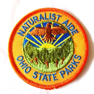 May include: Embroidered patch with the words "NATURALIST AIDE" and "OHIO STATE PARKS" in blue on a yellow background. The patch features a brown bird, green trees, and a blue sky with a yellow sun.