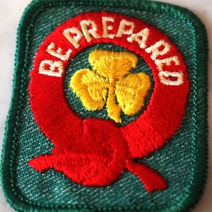 May include: Green fabric patch with a red embroidered circle around a yellow embroidered shamrock. The circle has the words "Be Prepared" embroidered in white.