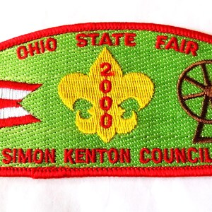 May include: Embroidered patch with a green background and red border. The patch features the text "OHIO STATE FAIR" in red, a yellow fleur-de-lis with "2000" in red, and a Ferris wheel. The Ohio state flag is on the left, and the text "SIMON KENTON COUNCIL" is at the bottom.